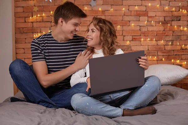 Sonriendo joven pareja hombre y mujer sentado en la cama y utilizando su ordenador portátil — Foto de Stock