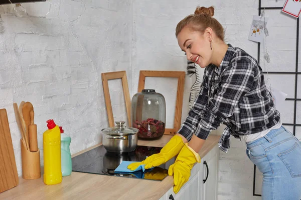 Mulher loira em luvas de proteção com pano de limpeza fogão elétrico em casa cozinha. Menina lavando superfície brilhante preto de cozinha superior — Fotografia de Stock