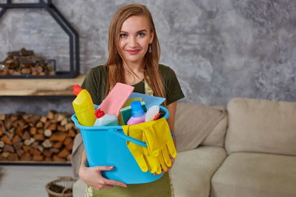 Una donna delle pulizie è in piedi in soggiorno con un secchio blu pieno di sostanze chimiche e strutture per riordinare in mano — Foto Stock