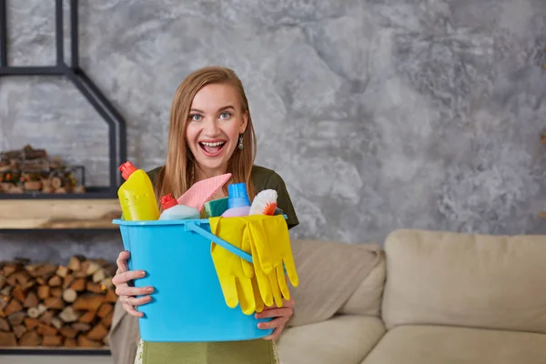 Uma mulher de limpeza está de pé na sala de estar segurando um balde azul preenchido com produtos químicos e instalações para arrumar em sua mão — Fotografia de Stock