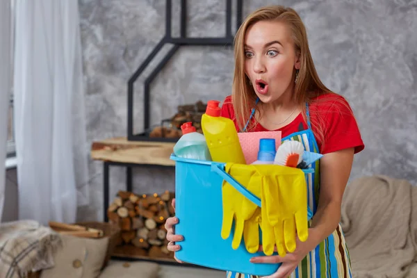 Una donna delle pulizie è in piedi in soggiorno con un secchio blu pieno di sostanze chimiche e strutture per riordinare in mano — Foto Stock