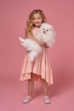 Pembe elbiseli sarışın bir kıvırcık saçlı küçük kız köpeğiyle oynuyor