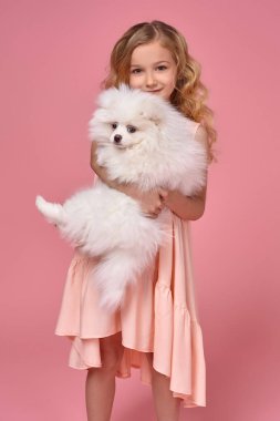 Pembe elbiseli sarışın bir kıvırcık saçlı küçük kız köpeğiyle oynuyor