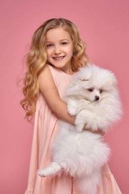 Pembe elbiseli sarışın bir kıvırcık saçlı küçük kız köpeğiyle oynuyor