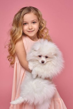 Pembe elbiseli sarışın bir kıvırcık saçlı küçük kız köpeğiyle oynuyor