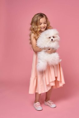 Pembe elbiseli sarışın bir kıvırcık saçlı küçük kız köpeğiyle oynuyor