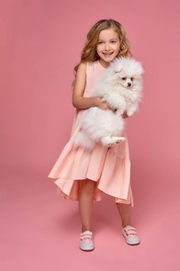 Pembe elbiseli sarışın bir kıvırcık saçlı küçük kız köpeğiyle oynuyor