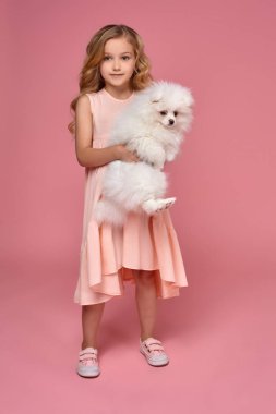 Pembe elbiseli sarışın bir kıvırcık saçlı küçük kız köpeğiyle oynuyor