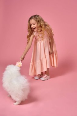Pembe elbiseli sarışın bir kıvırcık saçlı küçük kız köpeğiyle oynuyor