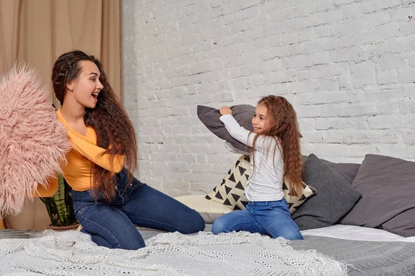 Young mom and daughter fooling around with pillow fights on the bed