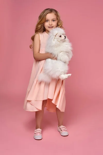 Egy szőke göndör haja, egy rózsaszín ruhás kislány játszik a kutyájával — Stock Fotó