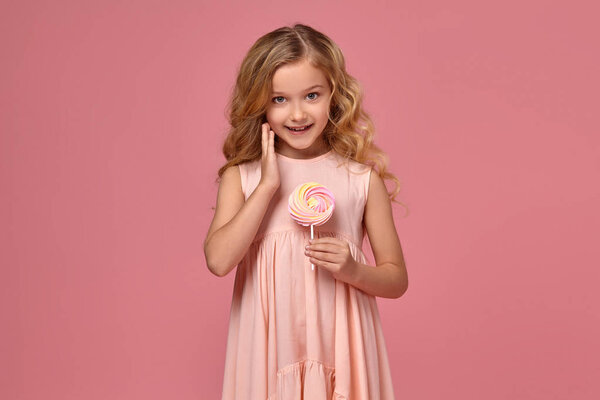 Маленькая девочка с кудрявыми светлыми волосами, в розовом платье позирует с конфетами
