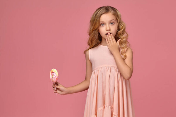 Маленькая девочка с кудрявыми светлыми волосами, в розовом платье позирует с конфетами

