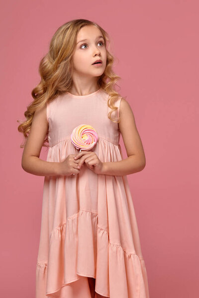 Маленькая девочка с кудрявыми светлыми волосами, в розовом платье позирует с конфетами
