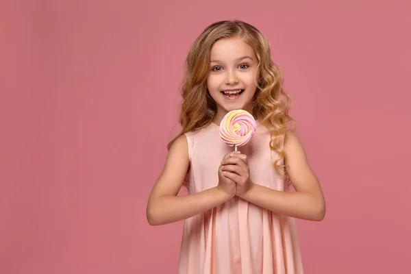 Ragazzina dai capelli biondi ricci, in abito rosa posa con una caramella — Foto Stock