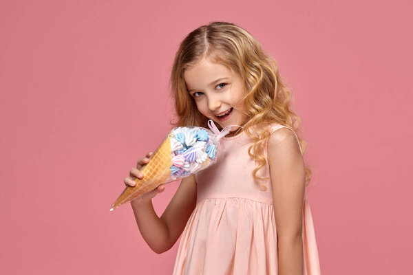Маленькая девочка с кудрявыми светлыми волосами, в розовом платье позирует с мороженым
