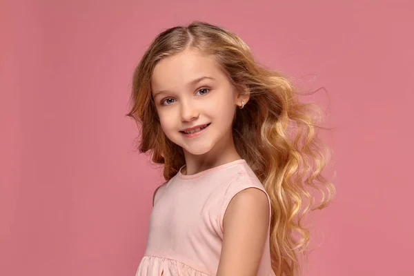 Pembe elbiseli sarışın bir kıvırcık saçlı küçük kız kamera için poz — Stok fotoğraf