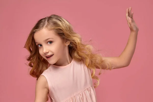 美丽的小女孩与金色卷发 在粉红色的礼服姿势为相机和看起来纳闷 在粉红色的背景 — 图库照片