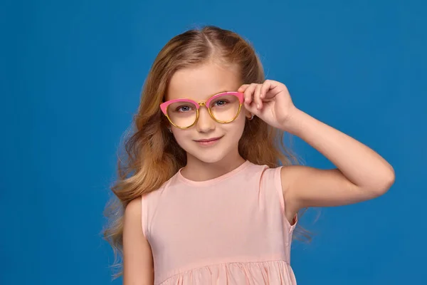 Módní malá holčička v růžových šatech a brýle stojí na modrém pozadí. — Stock fotografie