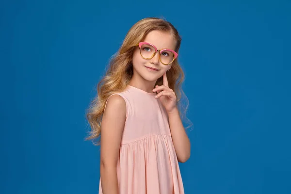 Szemüveg és egy rózsaszín ruha divatos kislány áll, kék háttérrel. — Stock Fotó
