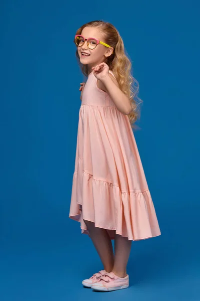 Niña de moda en un vestido rosa y gafas está de pie sobre un fondo azul . —  Fotos de Stock