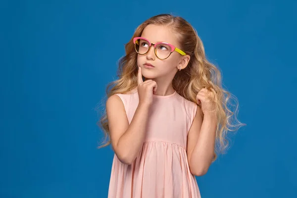 Μοντέρνο μικρό κορίτσι σε ένα ροζ φόρεμα και γυαλιά στέκεται πάνω σε μπλε φόντο. — Φωτογραφία Αρχείου