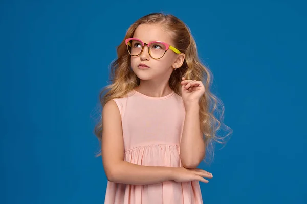 Modisches kleines Mädchen in rosa Kleid und Brille steht auf blauem Hintergrund. — Stockfoto