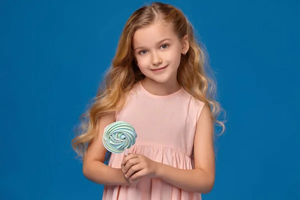 穿着粉色连衣裙的时髦小女孩, 手里拿着糖果, 站在蓝色的背景上. — 图库照片