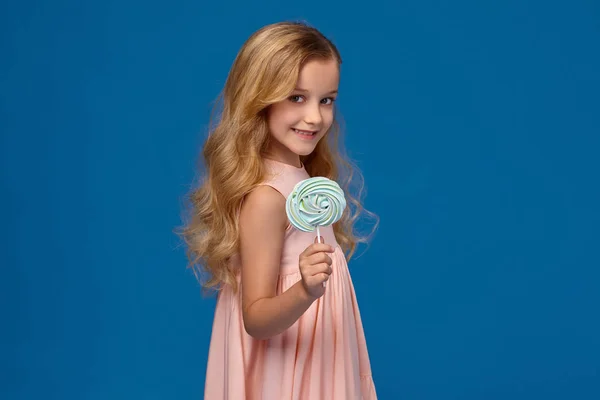 Niña de moda en un vestido rosa, con un caramelo en las manos, de pie sobre un fondo azul . — Foto de Stock