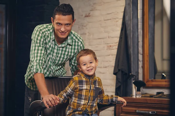 Garoto elegante sentado na cadeira na barbearia com seu jovem pai no fundo — Fotografia de Stock