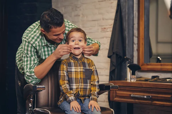 Garoto elegante sentado na cadeira na barbearia com seu jovem pai no fundo — Fotografia de Stock