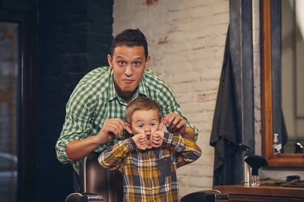 Stylisches kleines Kind sitzt auf Stuhl beim Friseur mit seinem jungen Vater im Hintergrund — Stockfoto