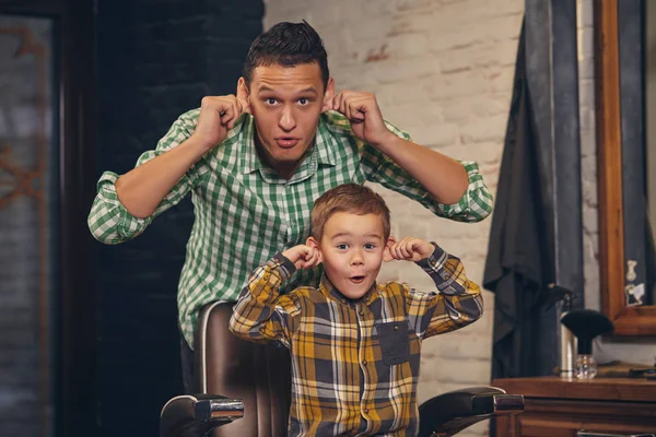 Stylish little kid sitting on chair at barbershop with his young father on background — Stock Photo, Image
