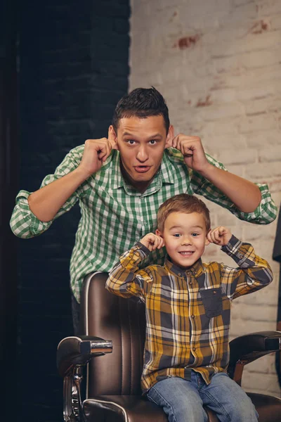 Стильна маленька дитина сидить на стільці в перукарні зі своїм молодим батьком на фоні — стокове фото