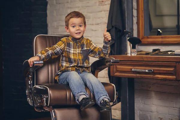 Portrét stylový malého chlapce oblečená v košili a džínách v holičství, sedí v křesle proti holiči pracoviště — Stock fotografie