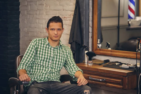 Schöner junger Mann schaut in die Kamera und sitzt auf einem Stuhl beim Friseur — Stockfoto