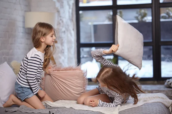 Gambar dua adik perempuan duduk di tempat tidur di kamar. Mereka menggunakan bantal untuk berkelahi satu sama lain — Stok Foto