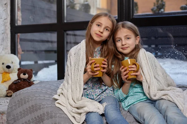 Изображение двух милых сестричек, сидящих на подоконнике, покрытых одеялом. Они пьют теплый чай. — стоковое фото