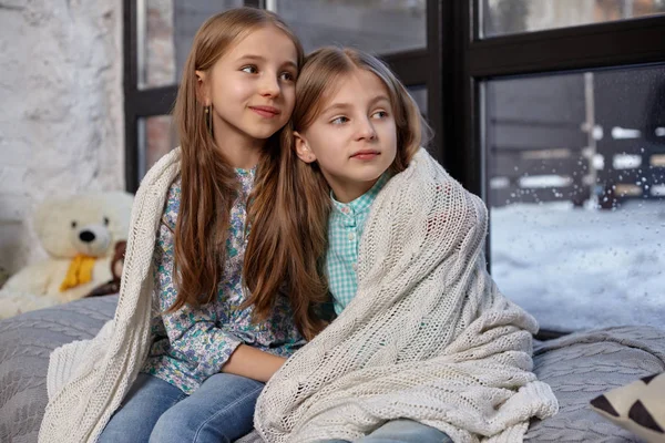 Изображение двух милых сестричек, сидящих на подоконнике, покрытых одеялом, обнимающих друг друга — стоковое фото