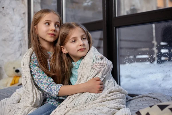 A imagem de duas irmãs pequenas bonitos sentados no peitoril da janela, cobertos por cobertor, abraçando um ao outro — Fotografia de Stock