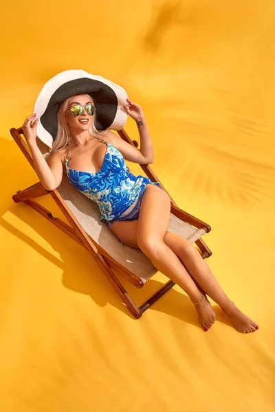 Beautiful young blonde woman in blue swimsuit lies on the lounger on yellow background — Stock Photo, Image
