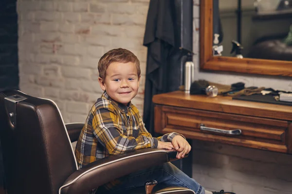 Retrato de un niño elegante vestido con camisa y jeans en la barbería, sentado en una silla contra el lugar de trabajo de los peluqueros — Foto de Stock