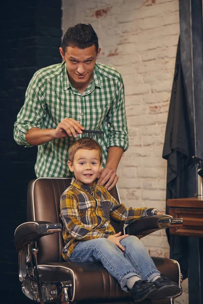 Stylisches kleines Kind sitzt auf Stuhl beim Friseur mit seinem jungen Vater im Hintergrund — Stockfoto