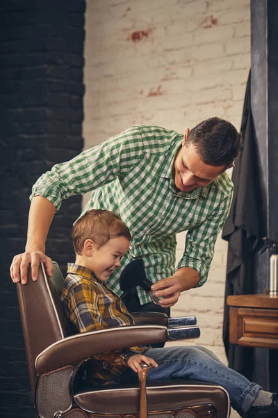 Garoto elegante sentado na cadeira na barbearia com seu jovem pai no fundo — Fotografia de Stock