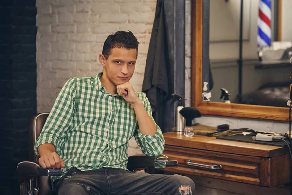Schöner junger Mann schaut in die Kamera und sitzt auf einem Stuhl beim Friseur — Stockfoto