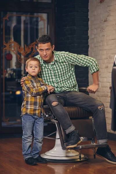 Mladý pohledný otec a jeho malý stylový syn v holičství čeká na holiče — Stock fotografie