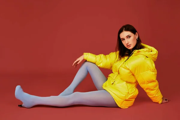 Retrato de estudio de una joven morena sentada en el suelo con chaqueta amarilla y pantalón azul gris mangueras o medias — Foto de Stock