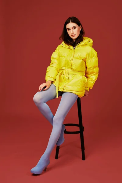 Hermosa joven mujer de moda en amarillo sobredimensionado chaqueta posando en el estudio sobre fondo rojo, utilizando taburete de bar — Foto de Stock