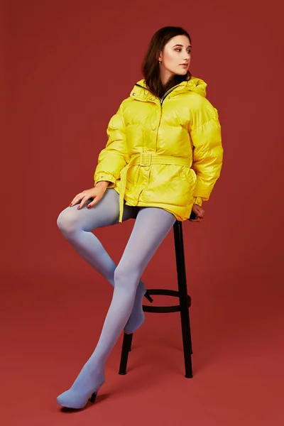 Hermosa joven mujer de moda en amarillo sobredimensionado chaqueta posando en el estudio sobre fondo rojo, utilizando taburete de bar — Foto de Stock