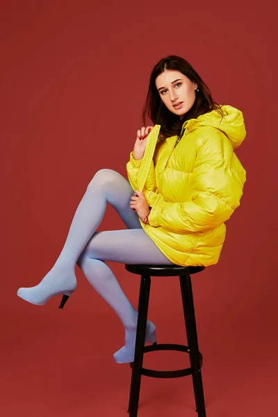 Bar tabure kullanan güzel genç moda kadın sarı aşağı büyük boy ceketli Studio kırmızı zemin üzerine poz — Stok fotoğraf